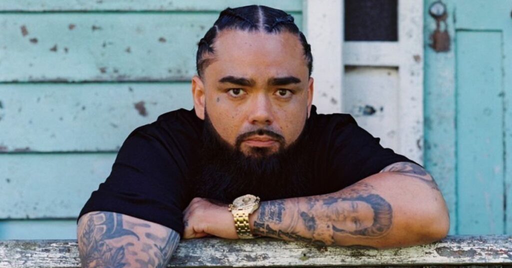 Poetik leaning on a wooden railing with tattoos and a gold watch, against a rustic teal background.